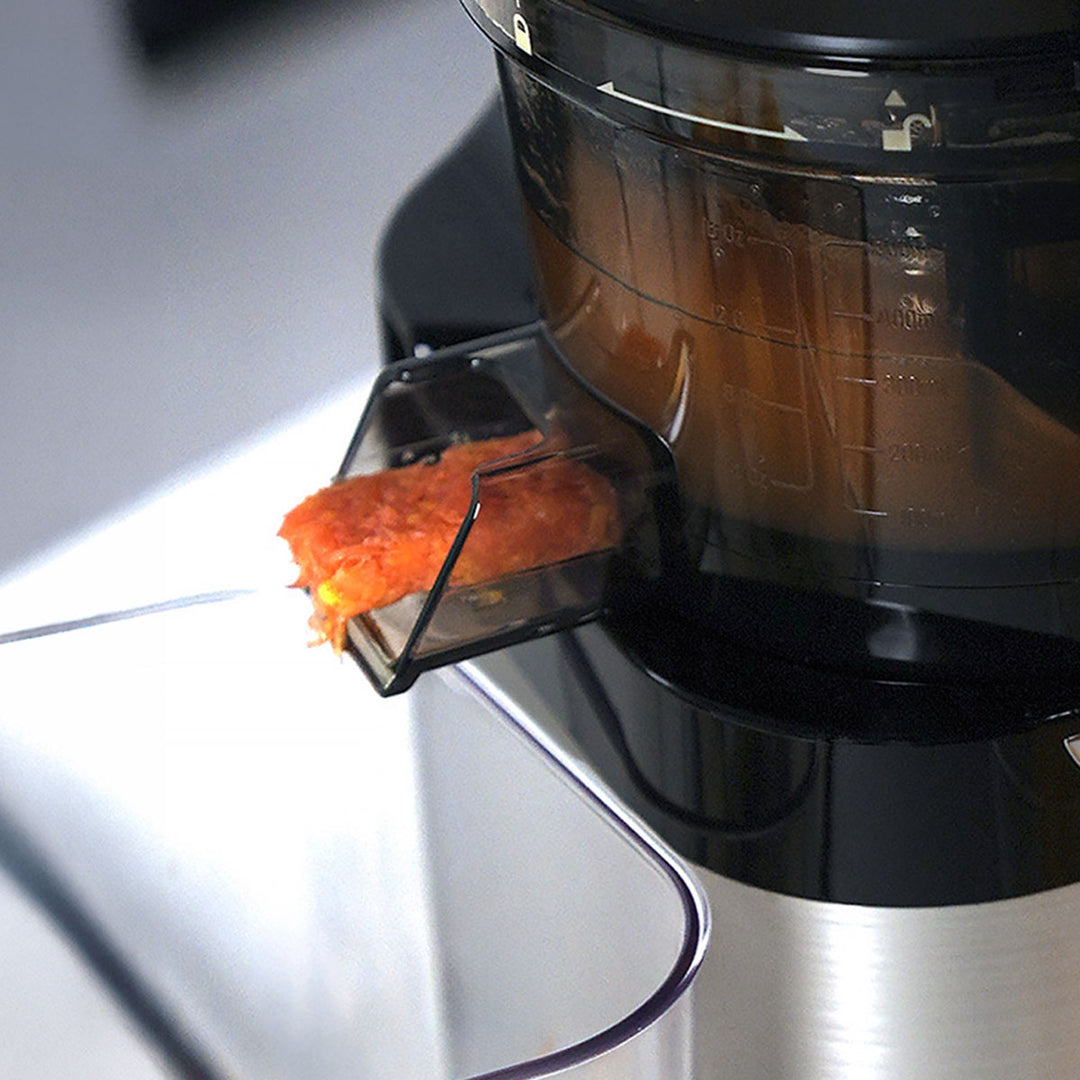 Close-up of a black and stainless steel countertop juicer with a clear feed tray holding orange juice pulp, with the dark juice collecting inside the chamber.