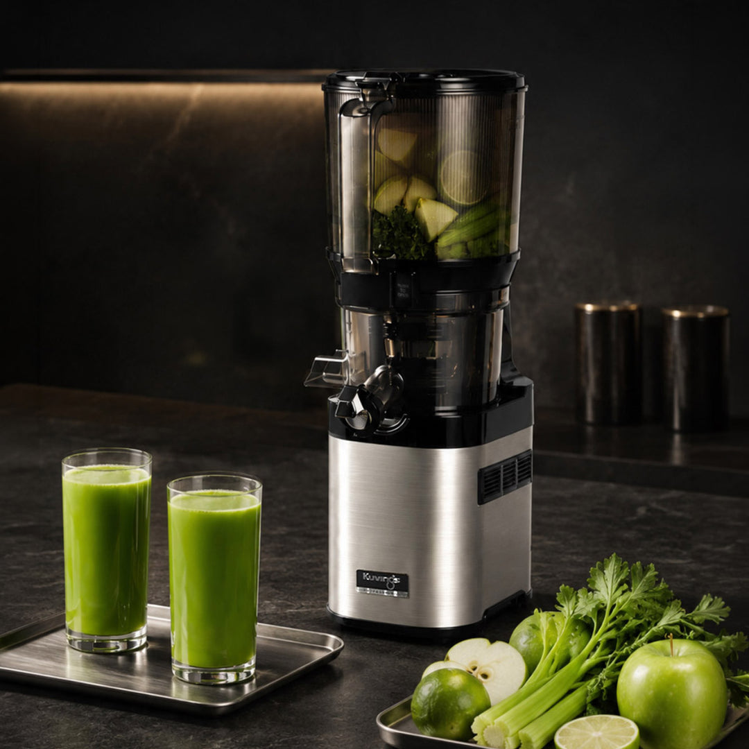 A countertop juicer machine with a clear upper chamber containing green juice and fruit, shown beside two glasses of green juice on a tray and fresh celery, apples, and limes on a dark kitchen counter.