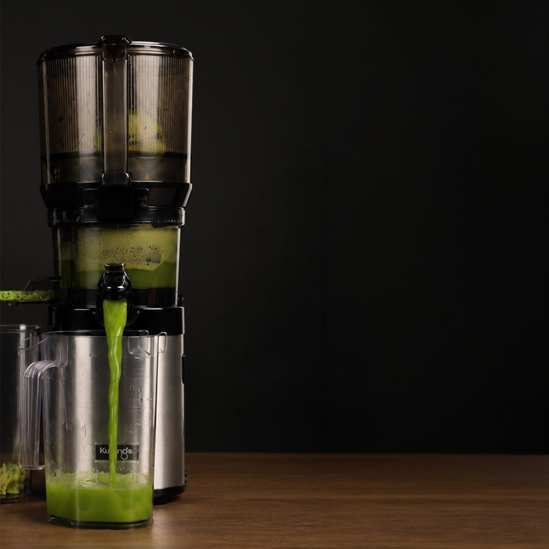 A countertop vertical Kuvings CS800 juicer with a clear and black body, shown dispensing bright green juice into a clear pitcher on a wooden surface against a dark background.