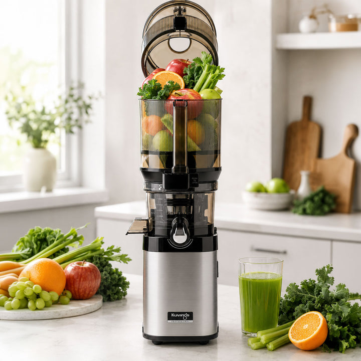 A countertop kitchen scene showing a stainless steel and black Kuvings slow juicer filled with mixed fruits and vegetables, with a glass of green juice and additional produce around it on a light stone countertop.