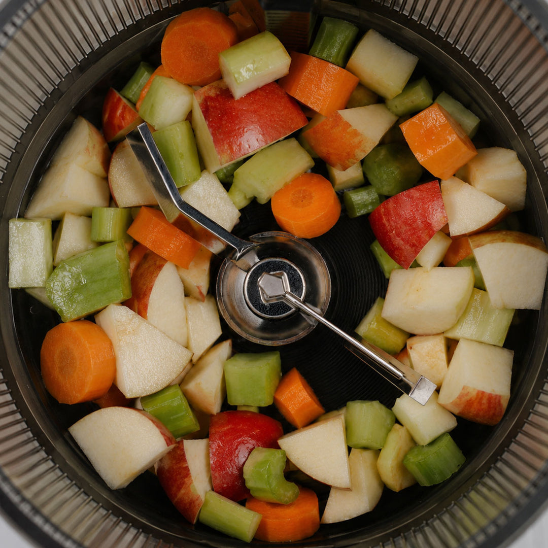 Chopped vegetables and fruit pieces inside a black food chopper bowl, including carrots, celery, apples, and pears, viewed from above.