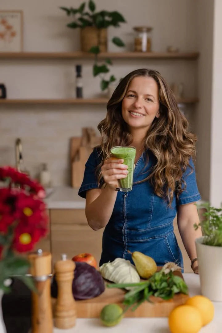 Zelené smoothie Kuvings držené ženou v kuchyni s čerstvým ovocem a zeleninou na pultu pro zdravé domácí recepty na smoothie.