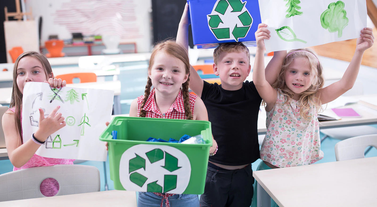 Group of elementary school children smiling in a classroom while holding up green and blue recycling bins and environmental drawings for an eco-friendly educational project.