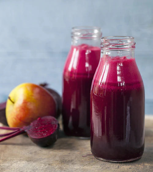 Zwei Flaschen Rote-Bete-Saft mit einem Hintergrund aus Äpfeln und Roter Bete.