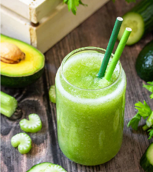 Grüner Smoothie in einem Glas mit Strohhalm, umgeben von frischem Gemüse auf Holzoberfläche