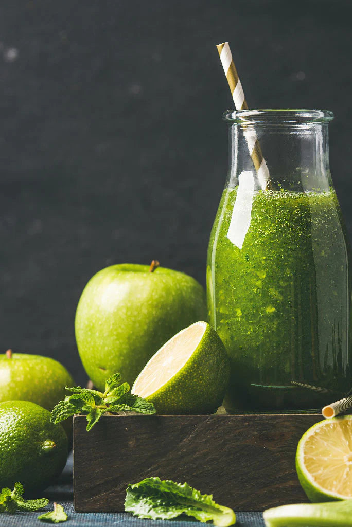 Glasflasche mit grünem Smoothie und Strohhalm, umgeben von grünen Äpfeln und Limetten auf dunklem Hintergrund