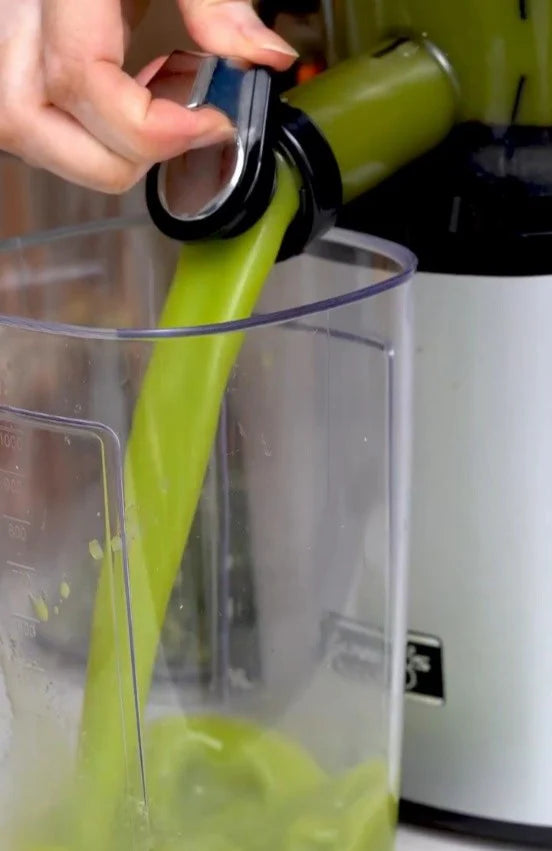 Zelené smoothie Kuvings držené ženou v kuchyni s čerstvým ovocem a zeleninou na pultu pro recepty na zdravé domácí smoothies.